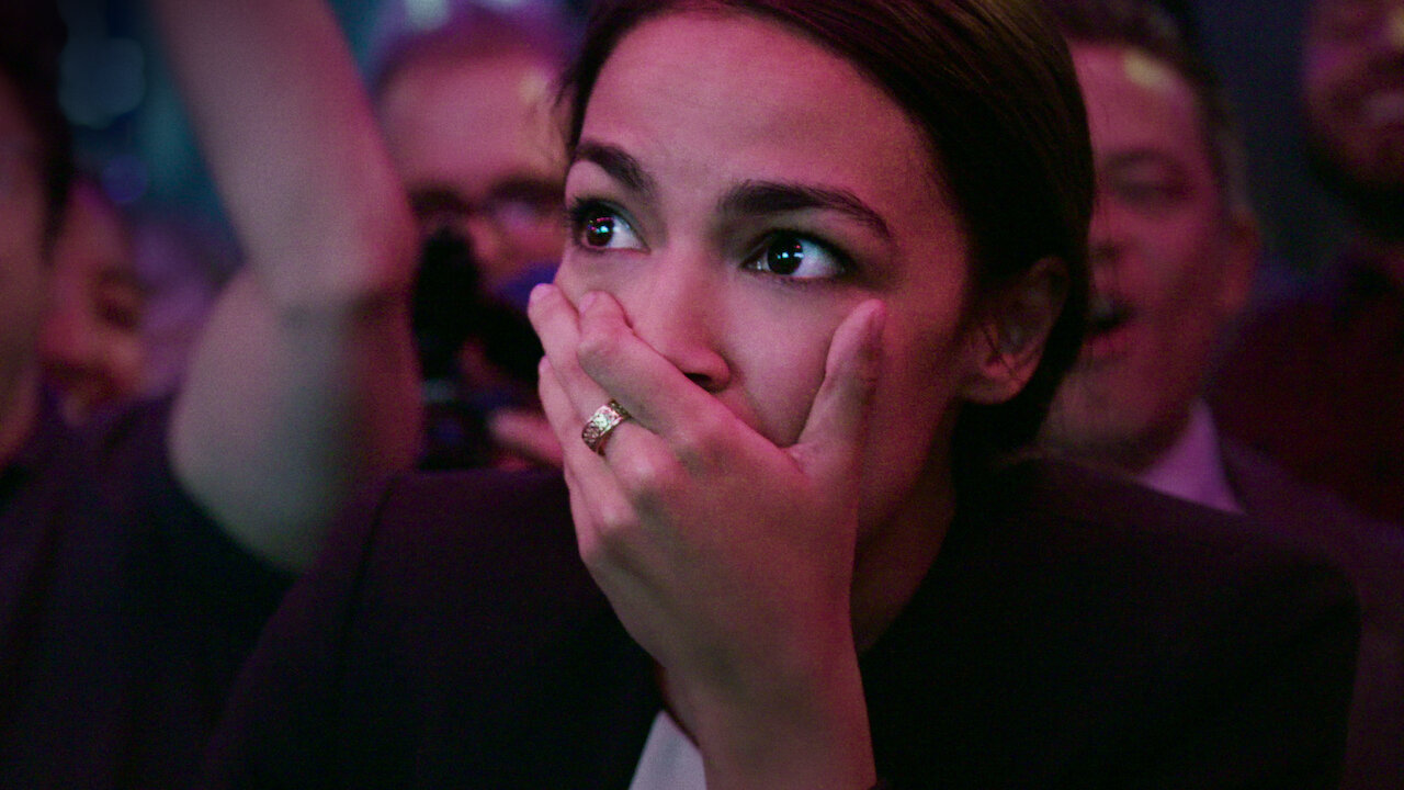 Image is from the documentary 'Knock Down the House' (2019). The image is of the politician Alexandria Ocasio-Cortez, who has her hands over her mouth after hearing she had won her campaign.